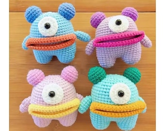 May include: Four colorful crochet monster toys. Each has a single eye, small arms, and a wide mouth. The monsters are in shades of blue, purple, pink, and green, with contrasting mouth and ear colors. These handmade toys are perfect for children.