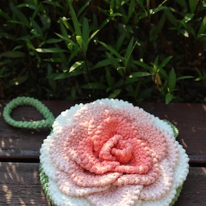 May include: A handmade, crocheted flower-shaped pillow in shades of pink, white, and green. The pillow has a spiral design, with a green handle attached. The pillow is sitting on a wooden surface, with green foliage in the background.