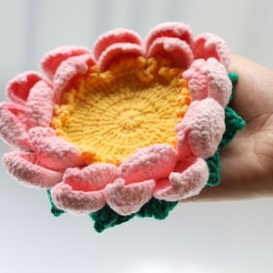 以下が含まれることがあります： ピンクの花びら、黄色の中心、緑の葉が付いた、手作りのかぎ針編みのフラワーコースター。 コースターは大きな花のように見えるように設計されています。 柔らかく、質感のある糸がユニークで装飾的なアイテムを作り出しています。