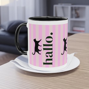 Op de afbeelding: Een keramische mok met een zwarte handgreep en binnenkant, voorzien van een verticaal patroon van lichtroze en witte strepen. Het ontwerp bevat zwarte silhouetten van katten en het woord "hallo." verticaal gedrukt in een vet, donkergroen schreeflettertype.