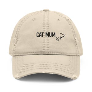 Könnte beinhalten: Eine beige Baseballkappe im Used-Look mit dem schwarzen Schriftzug CAT MUM und zwei kleinen schwarzen Herzumrissen. Die Kappe hat abgenutzte Kanten am Schirm und an den Vorderteilen, einen Knopf oben sowie gestickte Belüftungsösen.