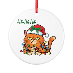 Könnte beinhalten: Ein rundes, weißes Keramikornament mit einem mürrischen, orangefarbenen getigerten Kater, der eine rote Weihnachtsmütze mit einer grünen Schleife trägt. Die Katze ist in bunte Lichterketten verwickelt, darüber steht der Text Ho Ho Ho in Grün.