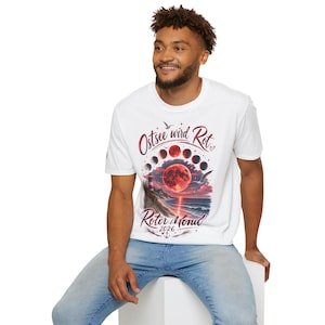 Könnte beinhalten: Ein weißes T-Shirt mit kurzen Ärmeln und einem Grafikdruck, der einen roten Mond über einer Küstenlandschaft mit Leuchtturm, Möwe und Mondphasen zeigt. Der Text lautet Ostsee wird Rot Roter Mond 2026 in roter Schrift.