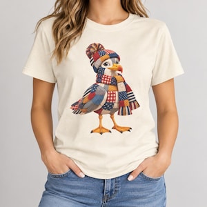 Könnte beinhalten: Ein cremefarbenes Kurzarm-T-Shirt mit einer Grafik eines Vogels, der eine Strickmütze und einen Schal trägt. Der Vogel hat ein Patchwork-Muster in Rot, Weiß und Blau sowie gelbe Füße und einen gelben Schnabel. Das Motiv ist vorne mittig platziert.