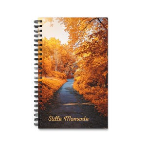 Könnte beinhalten: Ein spiralgebundenes Notizbuch mit einem Titelbild, das einen Weg durch einen Wald mit leuchtend orangefarbenem und gelbem Herbstlaub zeigt. Der Text 'Stille Momente' ist in goldener Schreibschrift unten auf dem Cover gedruckt.