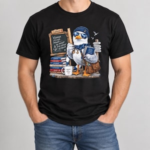 Puede incluir: Una camiseta negra con el gráfico de una gaviota que lleva un gorro azul, gafas de sol y una bufanda de lunares. La gaviota sostiene un libro y está junto a una pizarra con la palabra Lehrer, libros apilados y una taza con Beste Klasse.