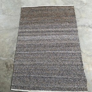 Puede incluir: Alfombra rectangular con superficie texturizada. La alfombra presenta una mezcla de fibras marrones, grises y blancas, creando una apariencia moteada. Los bordes están acabados con un borde blanco. La alfombra está diseñada para uso en interiores.