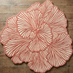 May include: A coral-coloured, flower-shaped rug with a cream-coloured centre. The rug features a detailed, textured design with petal-like patterns radiating outwards. The rug is placed on a wooden floor, showcasing its decorative appeal.
