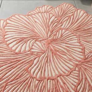 May include: A round rug with a floral design. The rug has a cream-coloured background with a raised, coral-coloured pattern. The pattern resembles a stylized flower with radiating lines and a scalloped edge.