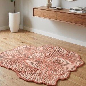 May include: A coral-coloured, floral-shaped rug with a cream-coloured centre. The rug features a detailed, raised pattern resembling leaves or petals. The rug is placed on a light-coloured wooden floor, adding a touch of nature-inspired decor to the room.