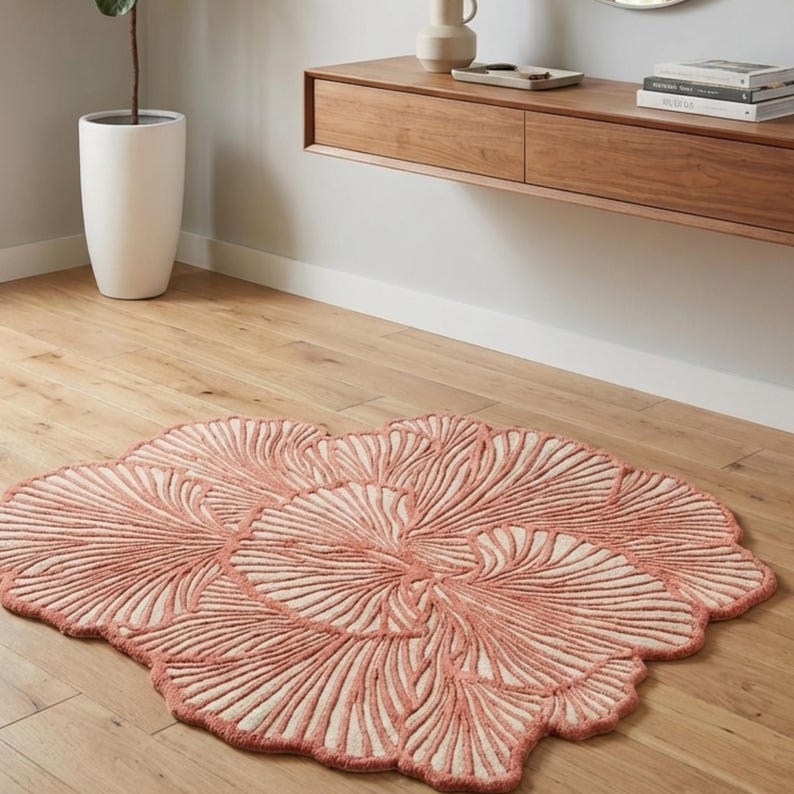May include: A coral-coloured, floral-shaped rug with a cream-coloured centre. The rug features a detailed, raised pattern resembling leaves or petals. The rug is placed on a light-coloured wooden floor, adding a touch of nature-inspired decor to the room.