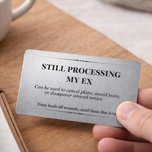 May include: A white ceramic mug with coffee sits on a wooden surface next to a card that reads "STILL PROCESSING MY EX." The card's text includes instructions for avoiding contact. A notebook and a tablet are also visible.