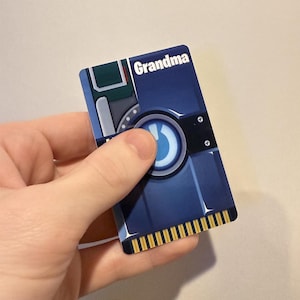 May include: A blue and gray card with the word "Grandma" in white text. The card has a circular design with a curved arrow and gold-colored connectors at the bottom. The card is held by a person.