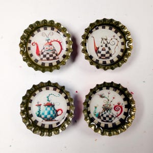 May include: Four bottle cap magnets with whimsical teapot designs. Each cap has a unique teapot illustration in red, blue, and black and white checkered patterns. The caps have a metallic, crimped edge.