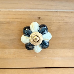 May include: A decorative cabinet knob with a flower design. The knob features alternating black, off-white, and gold petals. The center of the knob is a gold-colored, circular design. The knob is mounted on a light-colored wooden surface.