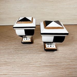 May include: Two black and white ceramic finials with gold accents. The finials are square with a pyramid-shaped top. They have a black and white checkered pattern with gold trim.
