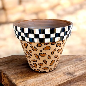 May include: A terracotta plant pot featuring a hand-painted leopard print pattern on the exterior body and a black and white checkered border around the upper rim. The interior of the pot is a smooth, unpainted brown clay finish.