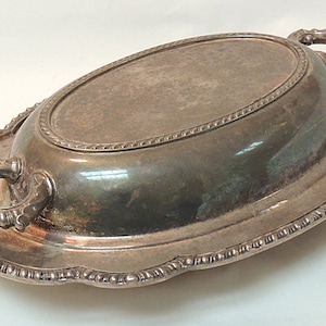 May include: Silver-toned oval serving dish with handles. The dish has a decorative rope-like border around the rim and a scalloped edge. The handles are ornate and add to the vintage aesthetic. A classic piece for serving food.