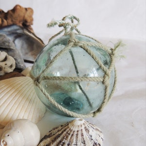 May include: A clear glass float, wrapped in a beige rope net, sits among seashells and driftwood. The float has a light blue-green tint and is tied with a rope at the top. The seashells are white and beige, creating a coastal theme.