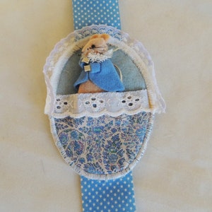 May include: A handmade Easter-themed bookmark featuring a felt rabbit in a blue coat, inside a floral patterned basket. The bookmark has a blue polka dot ribbon and is trimmed with white lace.