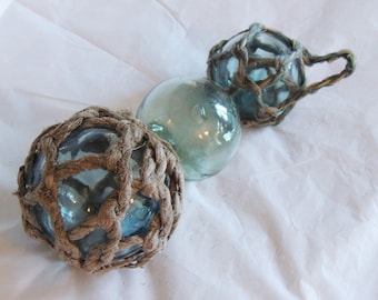 May include: Three glass fishing floats. Two are encased in woven rope netting, and one is a clear glass sphere. The floats have a light blue hue, and the rope is a natural brown color. The floats are arranged on a white surface.