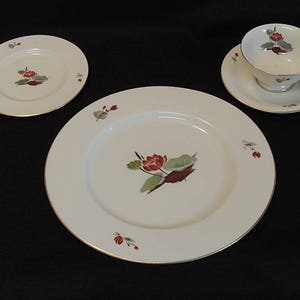 May include: A cream-colored porcelain tea set featuring a large plate, two smaller plates, a teacup, and a saucer. Each piece is decorated with a red and green floral design and gold trim. The set is arranged on a dark surface.