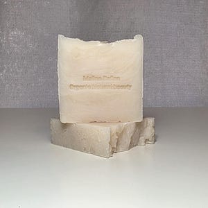 May include: Two bars of handmade soap, one stacked on top of the other. The top bar has the text "Malina Dalan Organic Natural Beauty" etched into it. The soap is a light beige color and has a textured, natural appearance.