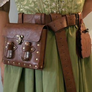 May include: A sage green corset with a white off-the-shoulder blouse, featuring a brown belt with a rectangular pouch containing two small glass vials, and a smaller leaf-shaped pouch attached to the side, all worn over a matching green skirt.
