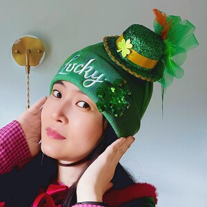 以下が含まれることがあります： 白い筆記体で「lucky」と書かれた緑色のニット帽。金色のバンドとクローバーが付いた、小さなキラキラ光る緑色のシルクハットが飾られています。緑のスパンコールのクラスターと、緑のチュールとオレンジ色の羽のアクセントが、お祝いのルックを完成させています。