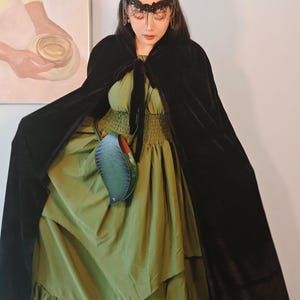 May include: A black velvet cloak with a tie closure, worn over a green dress with a ruffled hem. The dress has a fitted bodice and a flowing skirt. A decorative headpiece and a small green purse complete the ensemble.
