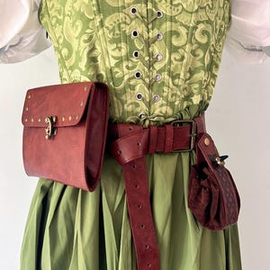 Puede incluir: Un corpiño verde con estampado, una blusa blanca debajo y una falda verde lisa. Un cinturón de cuero rojo oscuro se lleva en la cintura, con un bolso rectangular con solapa y tachuelas de latón y una bolsa pequeña con cordón ajustable.