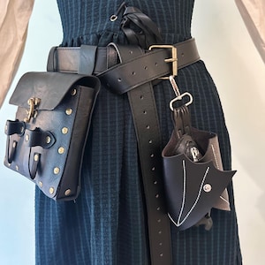 May include: A black leather belt featuring a large rectangular pouch with brass studs and a metal latch, alongside a smaller, wing-shaped leather pouch attached with a metal clip. The belt is worn over a dark green dress with a lace-up bodice.