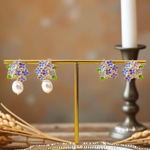 Könnte beinhalten: Blumenohrringe mit goldfarbenem Rahmen. Jeder Ohrring hat einen Cluster aus lila und blauen Blumen mit grünen Blättern und einer weißen Perle. Die Ohrringe werden auf einem goldenen Ständer präsentiert.