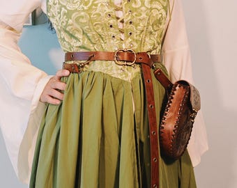 May include: A green and cream-colored medieval-style dress with a corset top and long skirt. The corset features a floral pattern and lace-up front. A brown leather belt and pouch complete the outfit, creating a historical or fantasy aesthetic.