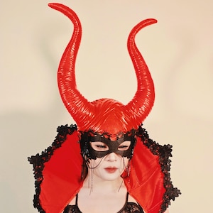 May include: A red devil horn headpiece with a matching red cape and black lace trim. The headpiece features large, curved horns and a black eye mask with dangling chains. The cape is trimmed with black lace.