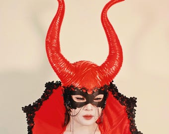 May include: A red devil horn headpiece with a matching red cape and black lace trim. The headpiece features large, curved horns and a black eye mask with dangling chains. The cape is trimmed with black lace.