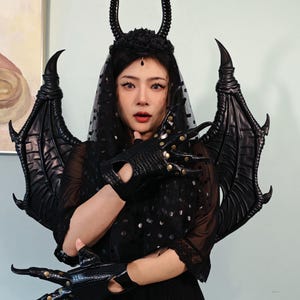 May include: A black costume set featuring a horned headpiece, bat-like wings, and clawed gloves. The headpiece has a floral base with curved horns. The gloves have long, pointed fingers with gold accents. The wings are large and detailed.