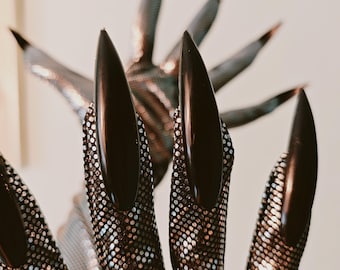 May include: A costume hand with long, black, glossy fingernails. The hand is covered in a silver mesh material with small, reflective dots. The background is a soft, neutral colour.