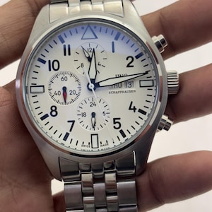 May include: A silver-tone stainless steel chronograph wristwatch with a white dial, black Arabic numerals, and a multi-link metal bracelet. The dial features three sub-dials, a day-date window at the three o'clock position, and the text IWC SCHAFFHAUSEN.