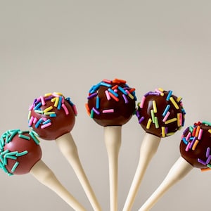 May include: Five chocolate cake pops with colourful sprinkles. Each round cake pop is dipped in dark chocolate and sits on a white stick. The sprinkles are a mix of blue, green, yellow, orange, and purple.