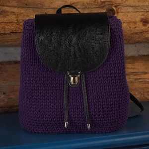 May include: A purple knitted backpack with a black leather flap and a silver clasp. The bag has a top handle and adjustable drawstring closures. The backpack is made of a thick, textured knitted material.