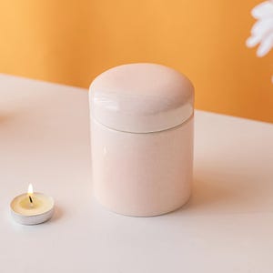 Puede incluir: Un pequeño tarro cilíndrico de cerámica rosa claro con tapa redondeada. Una vela de té encendida está a la izquierda. El fondo es naranja suave y blanco.