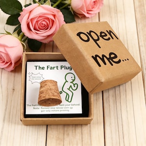 Op de afbeelding: Een novelty cadeau set met een kurken flesstopper met het label "The Fart Plug" in een kleine kartonnen doos. Op het deksel van de doos staat "open me..." in zwarte letters. Roze rozen zijn rond de doos gerangschikt op een houten oppervlak.