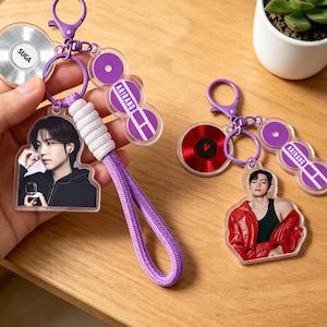May include: Two keychains with purple accents. One keychain features a photo of a person, a silver disc with the name "SUGA", and purple circles. The other keychain has a photo of a person, a red disc, and purple circles. Both have purple lanyards.