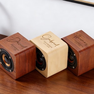 Può includere: Tre altoparlanti in legno personalizzati con nomi incisi sulla parte superiore. Gli altoparlanti sono a forma di cubo, con finitura in legno scuro su due e finitura in legno chiaro sull'altoparlante centrale. Ogni altoparlante ha un cono nero.
