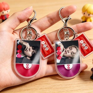 May include: Two keychains with a photo of a person, a red and a purple vinyl record design, and a red tag with white text. The keychains are held in a hand, with several small figurines in the background.