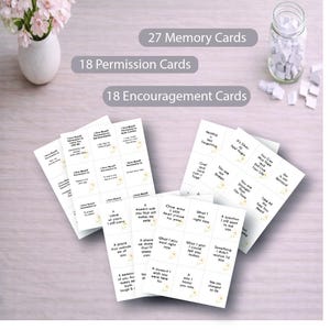 May include: A set of cards designed for reflection and support. The image displays 27 memory cards, 18 permission cards, and 18 encouragement cards. The cards are white with black text and a small floral design. A vase of pink flowers and a jar of small white papers are also present.