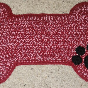 May include: Red and white crocheted dog bone shaped rug with a black paw print in the center.