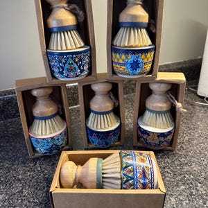 Puede incluir: Un conjunto de cepillos decorativos para lavar platos. Cada cepillo tiene un mango de madera, una banda azul y cerdas naturales. Las bases de cerámica están adornadas con coloridos diseños florales y geométricos, presentados en cajas individuales de cartón.