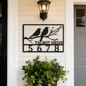 May include: Black metal address sign with two birds on a branch, the text "TALBOT DRIVE" and the numbers "5678". The sign is mounted on a white, horizontal-planked wall. A traditional outdoor lamp is above.